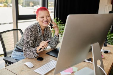 Neşeli kel kadın telefonda konuşuyor, yaratıcı bir alanda dizüstü bilgisayarıyla çalışırken gülümsüyor..