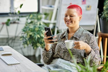 Canlı saçları olan kel bir kadın kahvesini yudumlarken gülümsüyor ve telefonunu parlak bir ofiste kontrol ediyor..