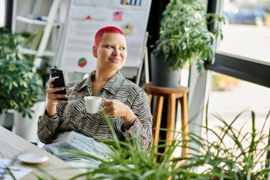Canlı saç stili olan kel bir kadın kahvesini yudumlarken gülümsüyor ve telefonunu içeride kontrol ediyor..