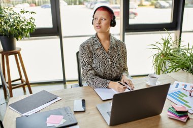 A bald woman with headphones is attentively writing notes at her desk surrounded by greenery. clipart