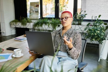 Seated at a modern desk, a bald woman utilizes her laptop while surrounded by plants. clipart