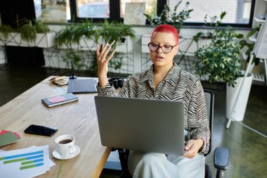 Şık gözlüklü kel bir kadın yemyeşil bir ortamda online bir toplantıya katılıyor..