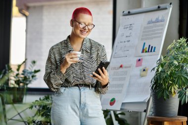 Şık giyinmiş kel bir kadın, yeşilliklerle çevrili telefonunu eleştirirken gülümsüyor..
