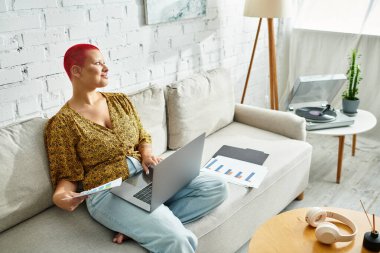 Kendine güvenen kel bir kadın dizüstü bilgisayarı üzerinde çalışırken gülümsüyor, etrafı kağıtlar ve dekorlarla çevrili..