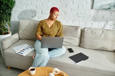 A stunning bald woman concentrates on her laptop while surrounded by a serene living space. clipart