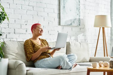 Kel kafalı neşeli bir kadın dizüstü bilgisayarıyla uğraşırken koltukta dinleniyor..