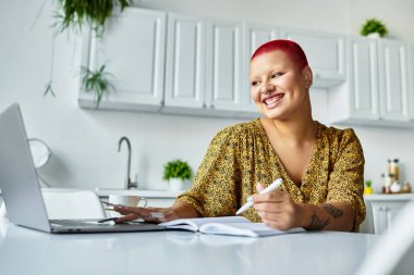 A woman with a radiant smile writes in a notebook while looking at her laptop in a modern kitchen. clipart