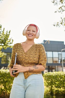 Neşeli, kel bir kadın elinde dizüstü bilgisayarla, kulaklık takmış, neşe saçıyor..