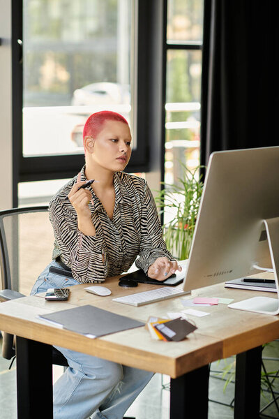 Женщина с яркой лысой головой созерцает свою работу, сидя за стильным столом.