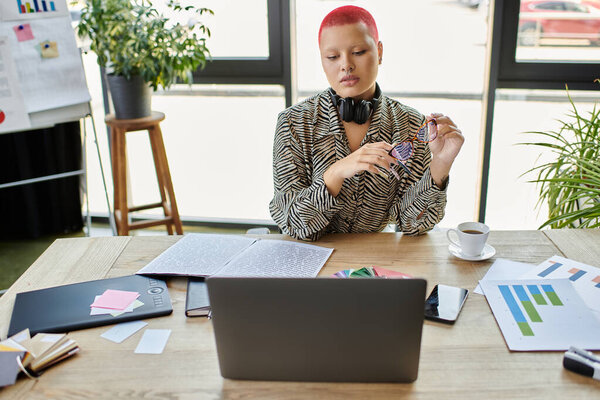 Модная лысая женщина сидит за своим столом и вдумчиво анализирует свои дизайны, сосредоточившись.