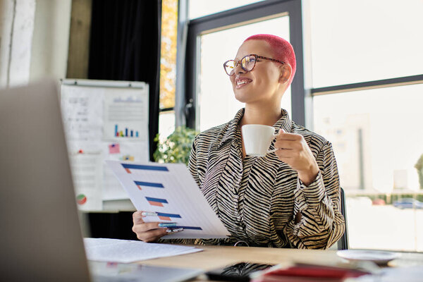 Радостная лысая женщина держит чашку кофе, анализируя отчеты на привлекательном рабочем месте.