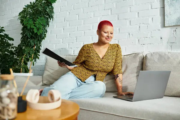 Kırmızı saçlı, kendine güvenen kel bir kadın şık bir evde dizüstü bilgisayarını kullanıyor..