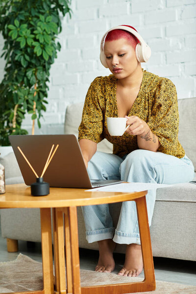 A calm setting features a bald woman with headphones sitting on a couch, sipping tea.