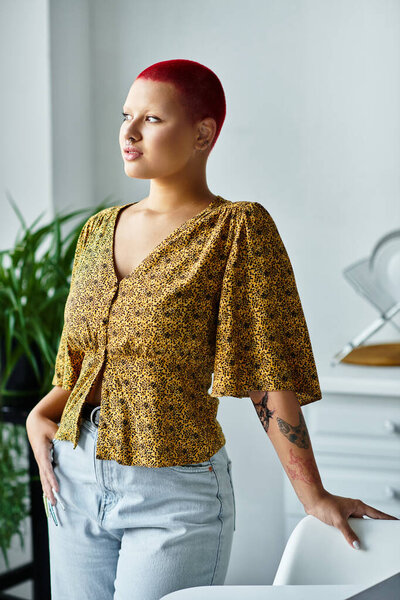 Лысая женщина со смелым красным цветом волос уверенно позирует в стильной, освещенной солнцем обстановке интерьера.