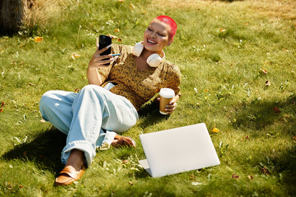 A cheerful bald woman lounges on the grass, sipping coffee and engaging on her phone, relaxed.
