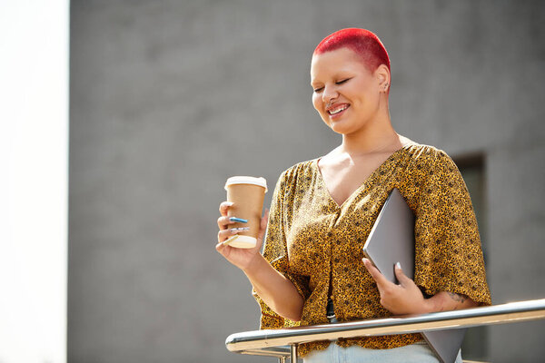 A woman with a bright red head is savoring her coffee and smiling while standing outside.