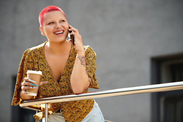 Светлая лысая женщина улыбается, держа в руках кофе и разговаривая по телефону на улице.