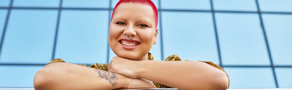 A bald woman with bold red hair stands smiling, showcasing her unique style outdoors.