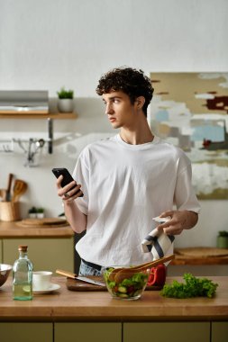 Yakışıklı genç bir adam şık bir mutfakta telefonunu kontrol ederken yaratıcı bir şekilde sağlıklı bir salata yapıyor..