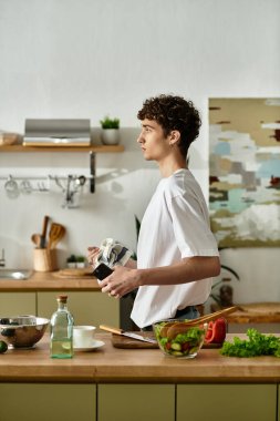 Kıvırcık saçlı yakışıklı genç bir adam şık bir mutfakta canlı bir salata hazırlıyor ve sağlıklı bir hayatı kucaklıyor..