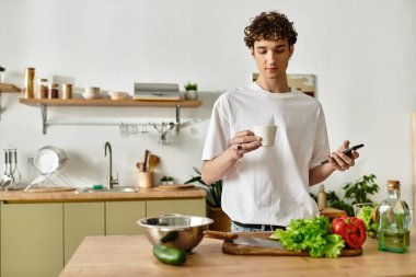 Yakışıklı genç bir adam elinde fincanla telefonuna bakar ve şeffaf bir mutfakta taze salata yapar..