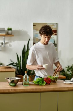Parlak bir mutfakta, kıvırcık saçlı genç bir adam sağlıklı bir salata için taze malzemeleri karıştırır..
