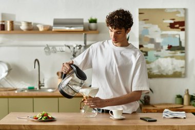 Çağdaş bir mutfakta, yakışıklı genç bir adam çaydanlıktan bardağa kahve doldurur, mutfak anlarından zevk alır..