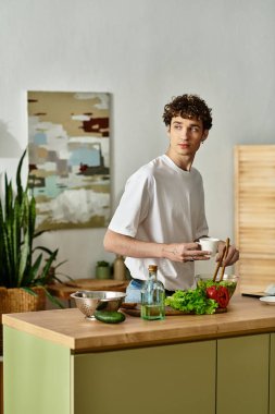 Şık bir mutfakta, kıvırcık saçlı genç bir adam neşeli bir şekilde renkli bir salata hazırlar, sağlıklı bir yaşam tarzını benimser..
