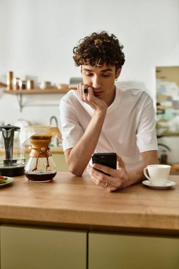 Sıcak bir mutfakta, kıvırcık saçlı yakışıklı genç bir adam telefonuna odaklanarak kahvesinin tadını çıkarıyor..