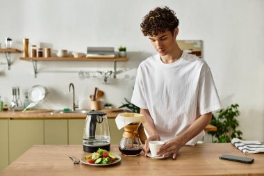 Yakışıklı genç bir adam sıcak bir mutfakta kahve hazırlıyor, elinde sıcak bir bardakla anın tadını çıkarıyor..