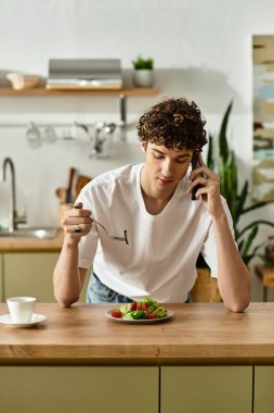 Modern bir mutfakta, genç bir adam sağlıklı bir tabak salata yerken telefonla konuşur..