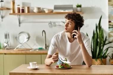 Yakışıklı genç adam renkli bir salatanın tadını çıkarıyor ve modern mutfağında telefon konuşmasından hoşlanıyor..