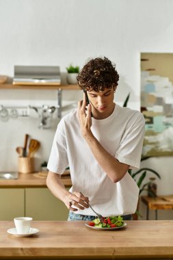 Parlak, şık bir mutfakta kıvırcık saçlı genç bir adam taze salatayı hazırlıyor ve telefonda sohbet ediyor..