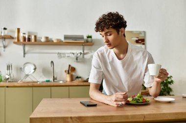 Kıvırcık saçlı yakışıklı genç adam modern, parlak bir mutfakta taze salata ve kahvenin tadını çıkarıyor..