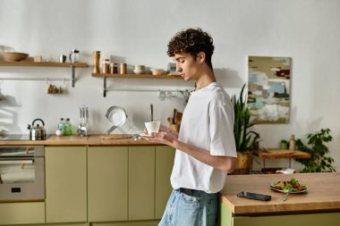 Yakışıklı genç bir adam çağdaş bir mutfakta duruyor, yanında renkli bir salata ile kahveyi yudumluyor..