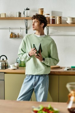 The handsome young man in a cozy kitchen contemplates while holding fresh green vegetables, exuding creativity. clipart