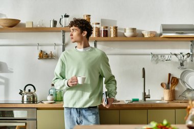 Düşünceli, kıvırcık saçlı genç bir adam şık bir mutfakta kahvesinin tadını çıkarıyor. Huzuru ve tarzı kucaklıyor..