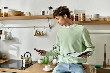 Parlak bir mutfakta, genç bir adam bir defter ve kahveyi dengelerken telefonuyla bir tarif üzerinde çalışır..