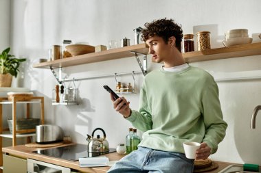 Şık bir mutfakta, yakışıklı genç bir adam tesadüfen kahvesini yudumluyor, akıllı telefonuna odaklanıyor..