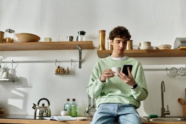 Yakışıklı genç bir adam şık bir mutfakta rahatlar, kahvesini yudumlar ve telefonunu rahat rahat karıştırır..