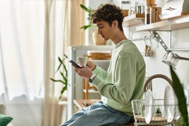 Yakışıklı genç bir adam kahvesini yudumluyor, telefonuyla modern bir mutfak ortamında meşgul..