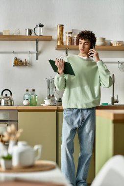 A stylish young man engages in a phone conversation while looking over a recipe in a chic kitchen setting. clipart