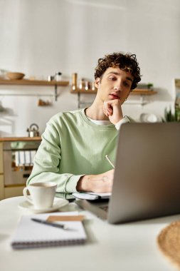 Yakışıklı bir genç adam dizüstü bilgisayarıyla çağdaş bir daireye odaklanırken düşünceli bir şekilde defterine yazar..