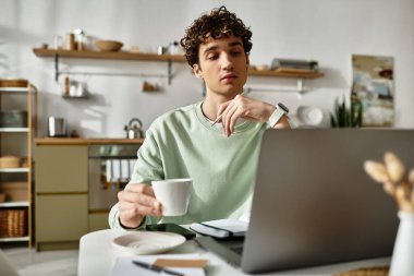 Kıvırcık saçlı bir adam, çağdaş bir ev ofisinde laptopuyla çalışırken dikkatlice kahvesini yudumluyor..