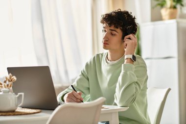 A young man thoughtfully writes notes while working remotely in his modern and bright apartment. clipart