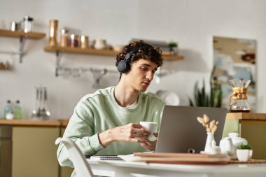 A focused young man works from home in a modern kitchen, sipping coffee while using his laptop. clipart