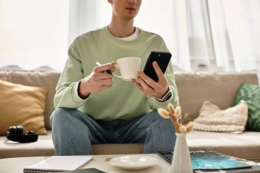 Genç bir adam rahat bir dairede akıllı telefonuyla uğraşırken kanepede dinleniyor, kahvesini yudumluyor..