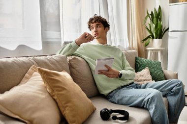 Kıvırcık saçlı genç bir adam evden çalışırken modern bir dairede düşünceli bir şekilde yansıtır..