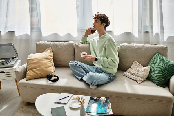 В яркой квартире красивый молодой человек пьет кофе и фокусируется на ноутбуке, расслабляясь на диване..