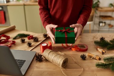 Genç bir adam evde Noel 'i kutlarken yeşil bir hediye kutusunu kırmızı kurdeleyle kaplıyor..